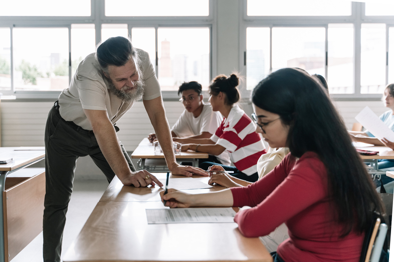 Friendly Professor explaining exercise to students in secondary school classroom. Senior Teacher with beard helping teenager learning student