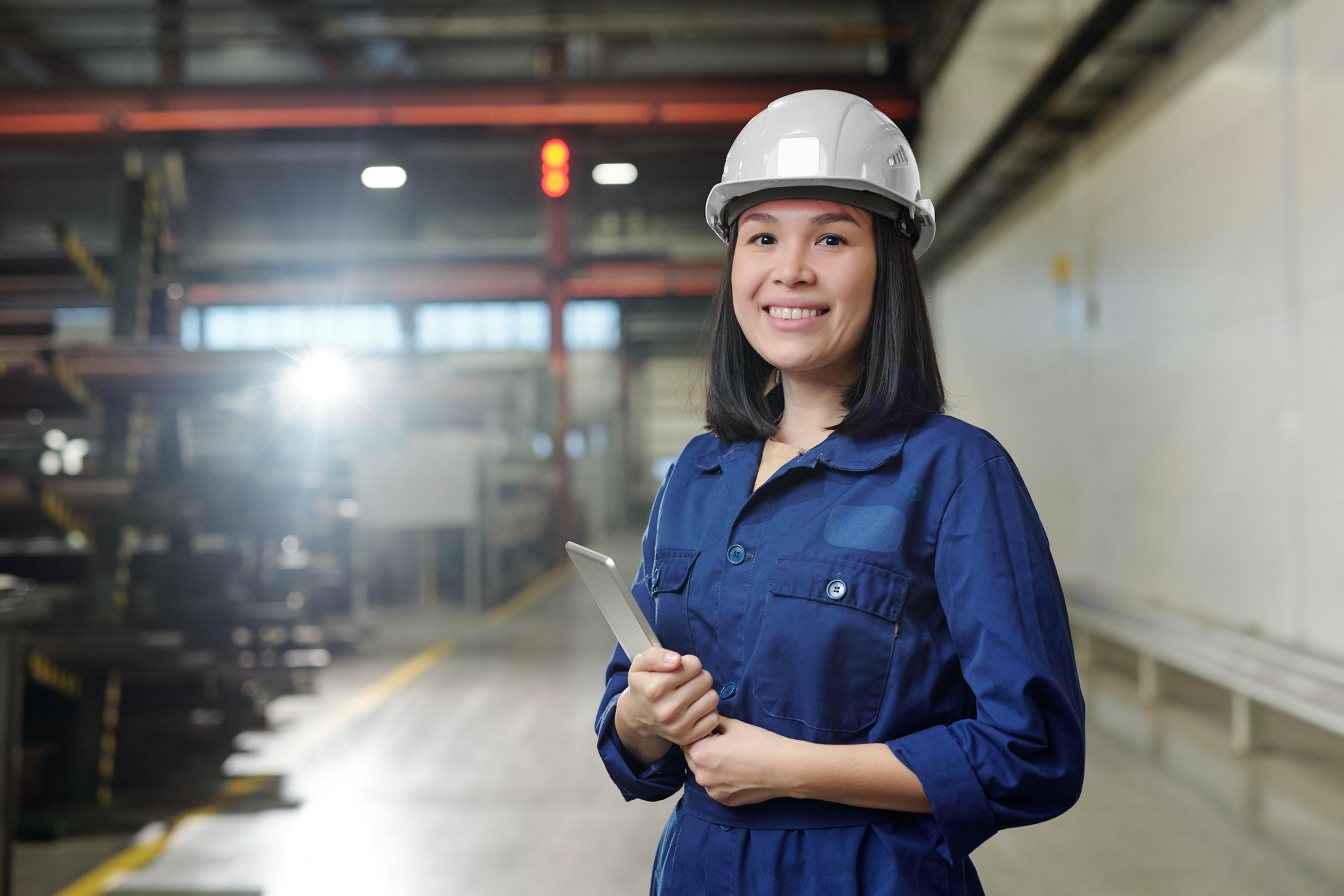 Pretty young smiling engineer in uniform and protective helmet holding tablet