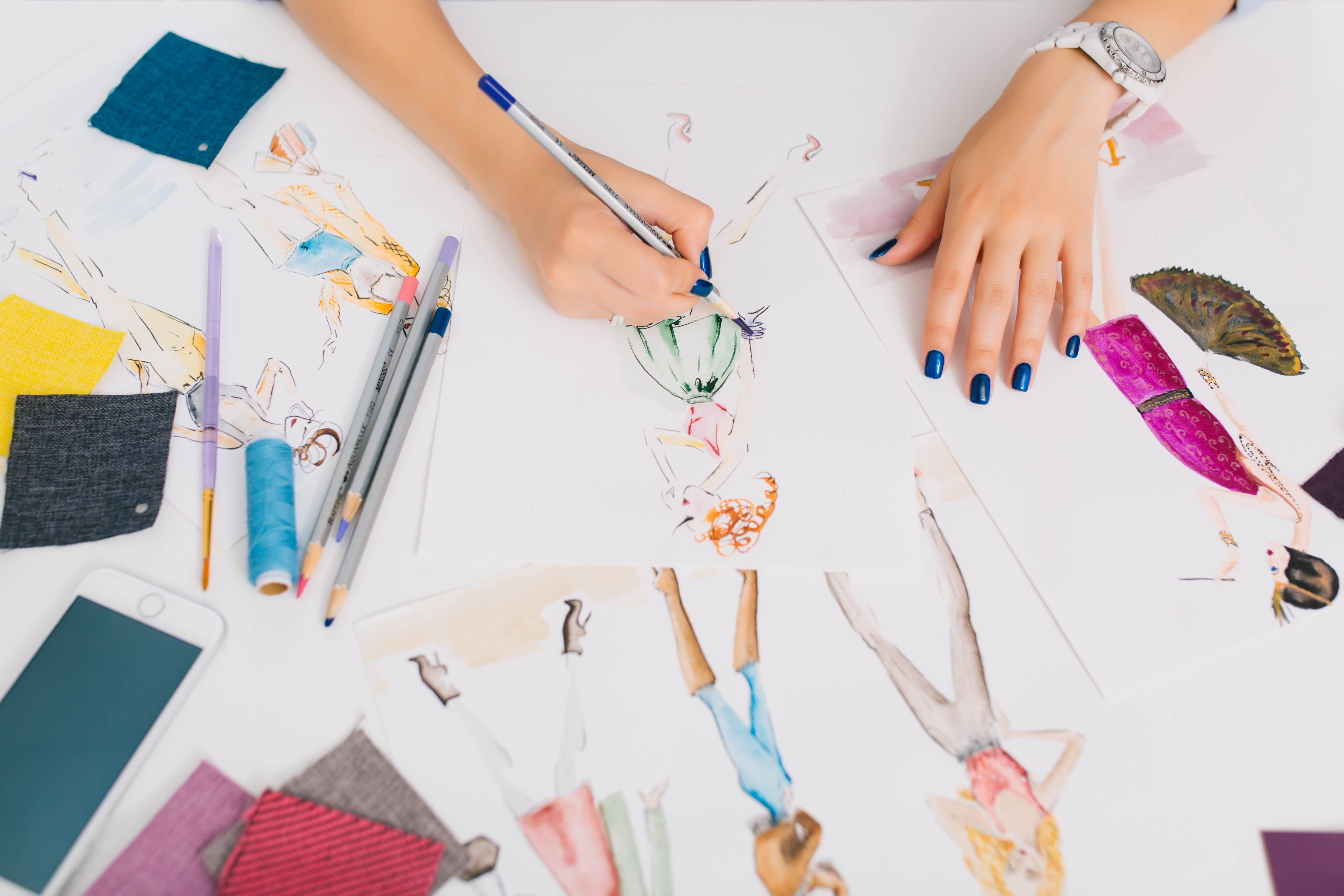 This picture describes the processes of designing clothes. There are hands of a girl drawing sketches on the table. There is creative mess with different stuff on the table
